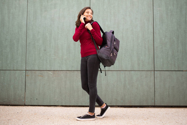 Full body profile portrait of young woman walking and talking with mobile phone and bag by wall - Photo, Image