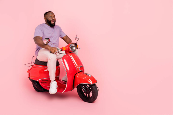 Photo of funny excited guy dressed violet t-shirt driving moped empty space isolated pink color background - Фото, изображение