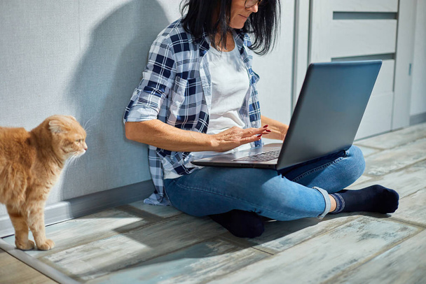 Volwassen vrouw zoekt dierenarts website op laptop te registreren kat voor veterinair overleg, Freelancer vrouwelijke typen op laptop zittend op de vloer thuis, werken vanuit huis. - Foto, afbeelding