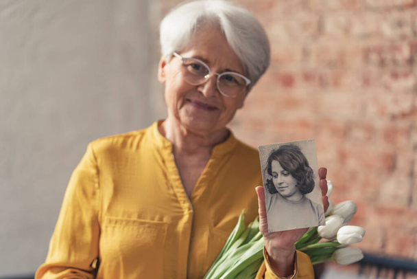 mujer mayor jubilada caucásica sosteniendo ramo de flores blancas y un retrato blanco y negro de su pasado - Foto, Imagen