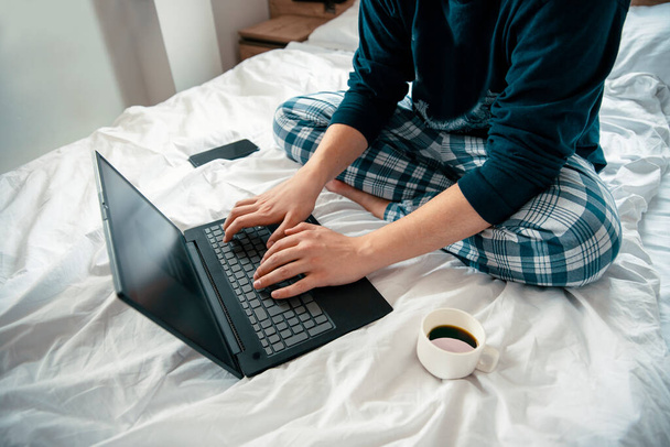 ベッドの中でノートパソコンを使って若い男のイメージを切り取る。Pajamaタイピングコンピュータキーボードの認識できない男性学生の上の側面図 - 写真・画像