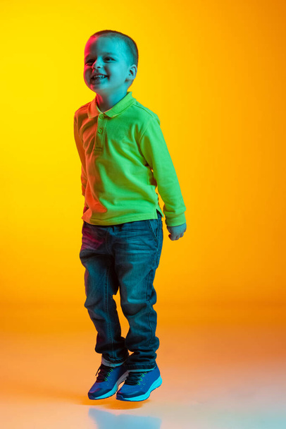 Full-length portrait of charming little boy posing isolated on yellow studio backgroud in neon light. Concept of child emotions, facial expression, action, childhood - Foto, immagini