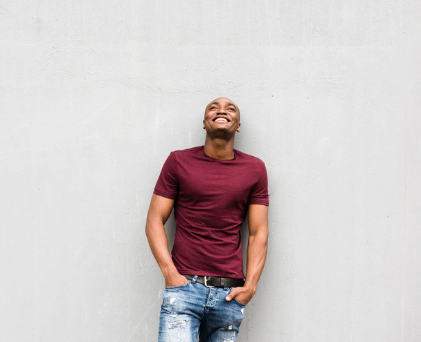 Portrait of happy young man standing by gray wall with hands in pockets and looking up - Photo, Image