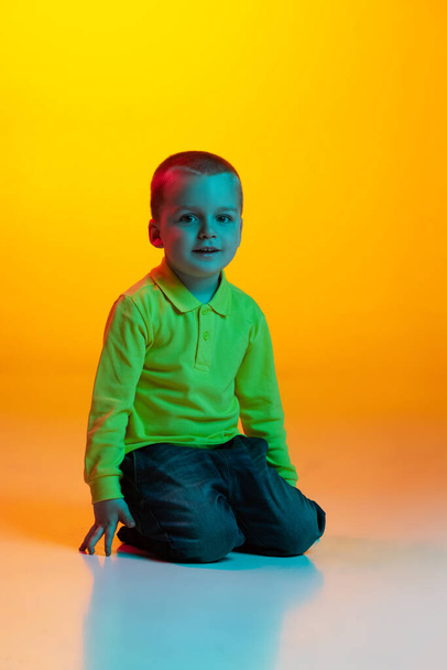 Studio shot of cute little boy, kid sitting on floor isolated on yellow studio backgroud in neon light. Concept of child emotions, facial expression, childhood - Foto, Imagem