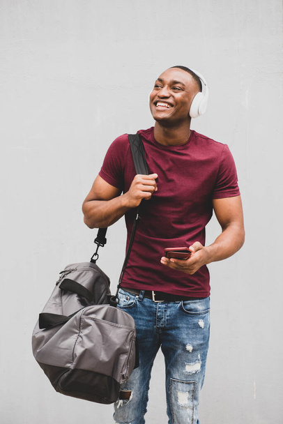 Portrét šťastného Afroameričana s taškou a mobilním telefonem, který poslouchá hudbu na sluchátkách  - Fotografie, Obrázek