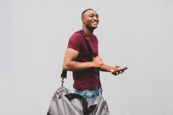 Profiel portret van gelukkige Afro-Amerikaanse man staan met tas en mobiele telefoon in de hand door grijze achtergrond - Foto, afbeelding