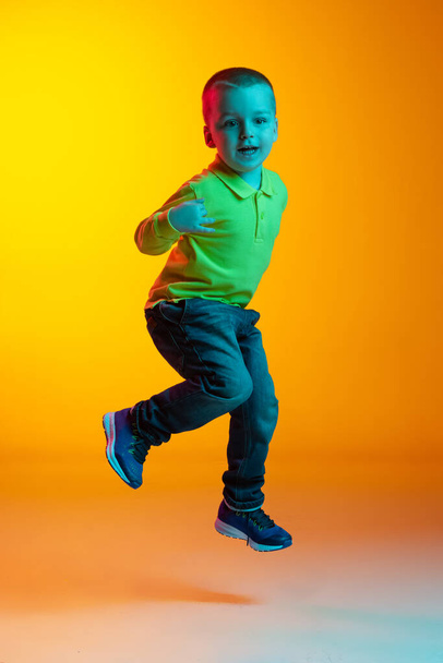Full-length portrait of charming little boy posing isolated on yellow studio backgroud in neon light. Concept of child emotions, facial expression, action, childhood - Foto, imagen