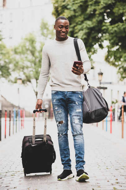 Πλήρες πορτρέτο του χαρούμενου Αφροαμερικανού ταξιδιώτη που περπατάει στο δρόμο με κινητό τηλέφωνο και βαλίτσα - Φωτογραφία, εικόνα