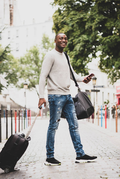 Ganzkörperporträt glücklich afroamerikanischen männlichen Reisenden zu Fuß auf der Straße mit Handy und Koffer - Foto, Bild