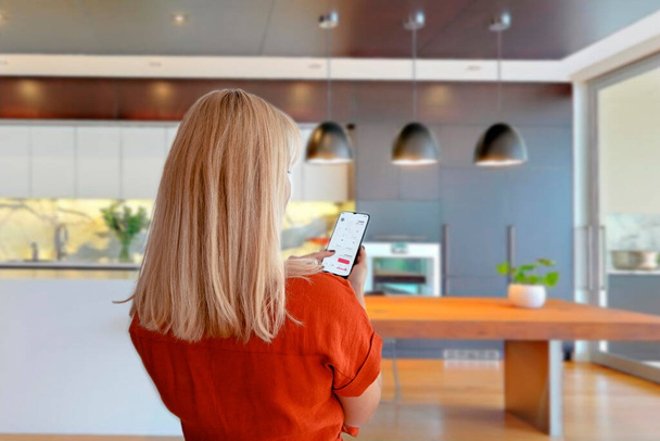 Nainen, joka käyttää älypuhelinsovellusta kotiautomaatioon. Blondi nainen ohjaa Smart kotiin mobiililaitteella - Valokuva, kuva