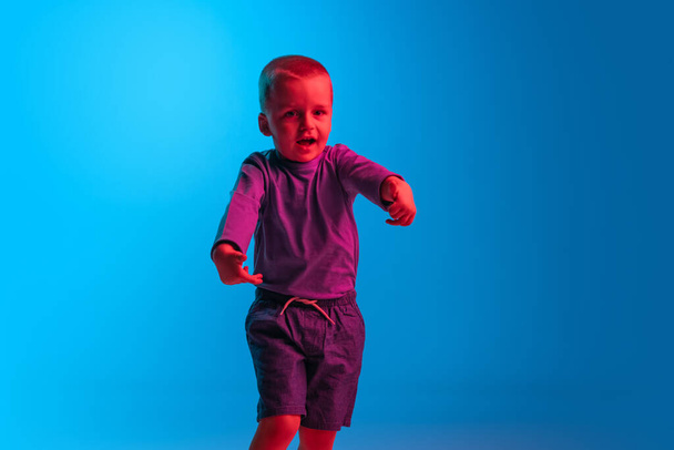 Portrait of little boy, kid, preschool age child isolated on blue studio backgroud in red neon light. Concept of child emotions, facial expression, childhood - Photo, Image