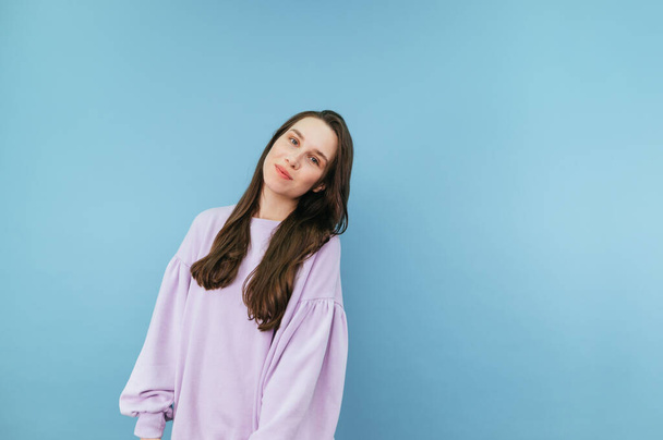 Beautiful brunette woman in a purple sweatshirt stands on a blue background and looks at the camera with a slight smile on his face. - Photo, Image