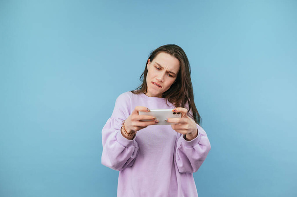 Focused brunette woman in purple sweatshirt playing online game on smartphone on blue background. - Photo, Image
