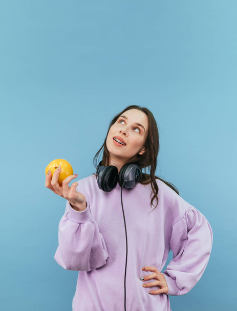 Pensiva donna carina in abiti casual si erge su uno sfondo blu con una mela tra le mani e le cuffie intorno al collo e guarda sognante - Foto, immagini