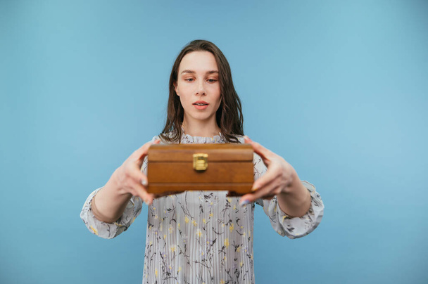 Όμορφη κυρία σε ένα φόρεμα στέκεται με ένα ξύλινο κουτί στα χέρια του σε ένα μπλε φόντο με ένα σοβαρό πρόσωπο κοιτάζοντας ένα κουτί κοσμήματα. - Φωτογραφία, εικόνα