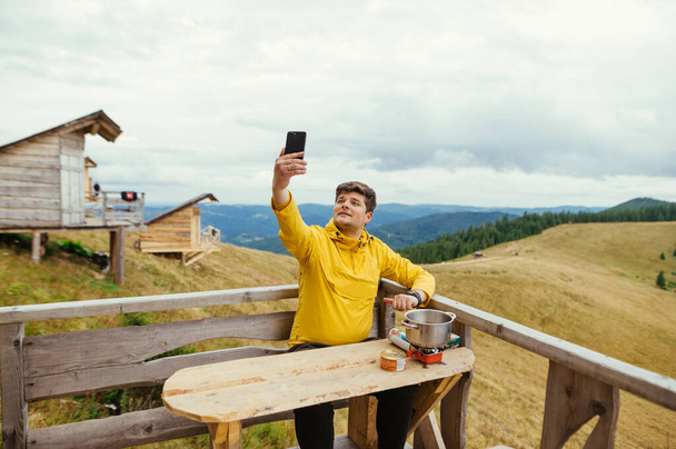 El hombre positivo está sentado en una terraza de bungalow en una mesa en las montañas, preparando comida y utilizando un teléfono inteligente con una cara alegre. - Foto, Imagen