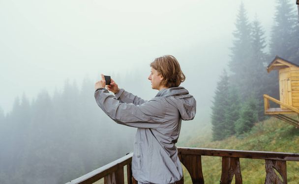 Mies turisti seisoo terassilla puutalo vuoristossa sumuisena aamuna ja ottaa kuvia kauniista maisemista älypuhelimen kameralla. - Valokuva, kuva