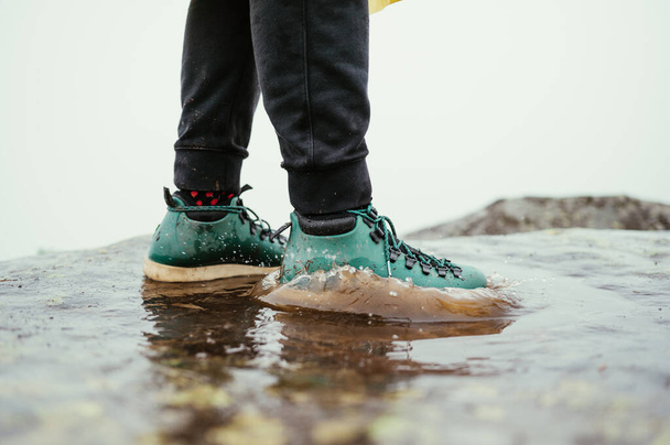 Πόδια στα παπούτσια ενός άντρα τουρίστα στέκονται σε μια λακκούβα σε ένα φόντο γκρίζων νεφών. - Φωτογραφία, εικόνα