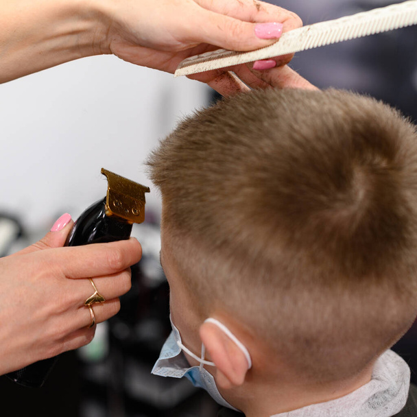 petit garçon dans un masque, qui est coupé dans le salon de coiffure dans le salon de coiffure, coupe de cheveux à la mode et élégant pour un enfant, machine à écrire de coupe de cheveux. - Photo, image