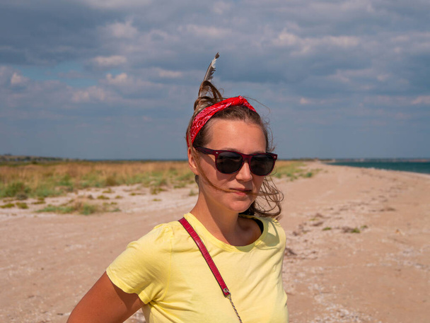 Fröhlich lächelnde junge Boho-Frau mit Feder im Haar am Strand. Modisches europäisches Mädchen in rotem Kopftuch und gelbem T-Shirt Millennial Generation Tribal Bohemian Style weibliches Porträt lange Haare - Foto, Bild