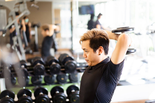 Active Asian sportsman doing a weight exercise in a gym - fitness by lifting up a dumbbell, man lifting up or holding a heavy dumbbell in Overhead Tricep Extension position. Bodybuilding workout. - Foto, Bild
