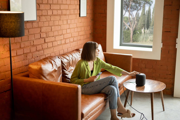 Frau berührt Smart-Lautsprecher, während sie zu Hause auf Couch sitzt - Foto, Bild