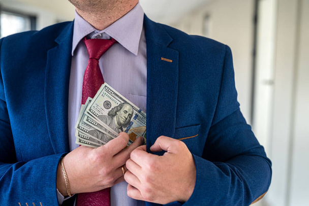 Facet w niebieskim garniturze i czerwonym krawacie wkładający do kieszeni banknoty łapówkowe. koncepcja korupcji  - Zdjęcie, obraz