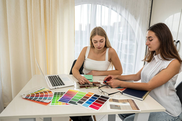 dos exquisitos trabajadores femeninos perfectos en blusas blancas trabajan en una mesa espaciosa en un nuevo diseño de habitación. Concepto de diseño - Foto, Imagen