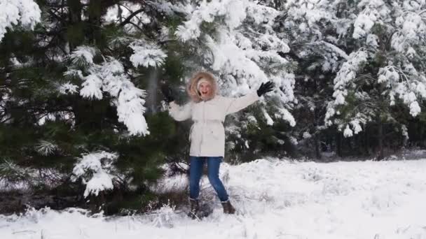 A woman in a white jacket in a winter pine forest jumps and has fun under snow-covered branches under falling snow - Filmagem, Vídeo