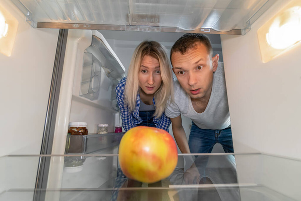 Un joven y una mujer miran una manzana en un refrigerador vacío. Concepto de servicio de entrega, pobreza. Foto desde el interior del refrigerador - Foto, imagen
