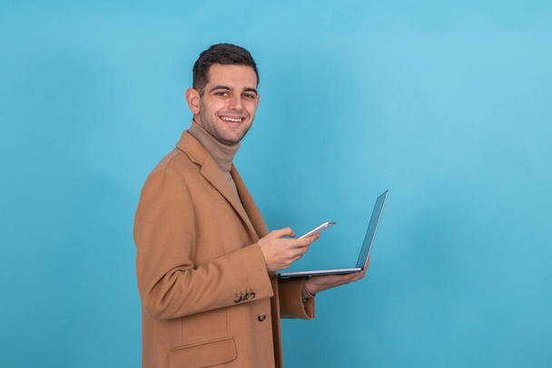 Mann mit Mantel, Handy und Computer isoliert auf blauem Hintergrund - Foto, Bild