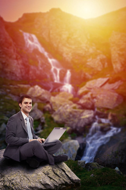 Businessman working on his laptop in the high mountains beside a waterfall at sunset, conceptual image of freedom - Фото, изображение