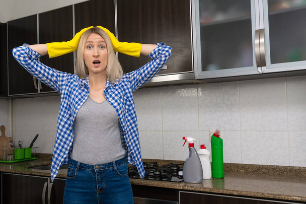 Mutfakta sarı eldivenli genç sarışın kadın başını tutuyor ve duygusal olarak yaklaşan temizlikten rahatsız oluyor ve tatmin olmuyor. - Fotoğraf, Görsel