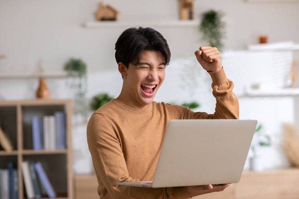 Asian man is smiling and expressing happy feeling on the computer laptop screen. young male got good news and show his cheerful face.Happiness men looking on laptop read message feel excited at home - Foto, afbeelding