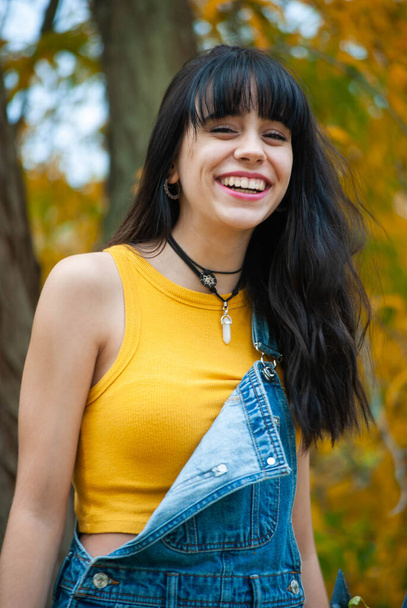 Nuori tyttö pukeutunut keltainen ja yllään denim haalarit hymyillen puistossa syksyllä vihreä ja keltainen väri. - Valokuva, kuva