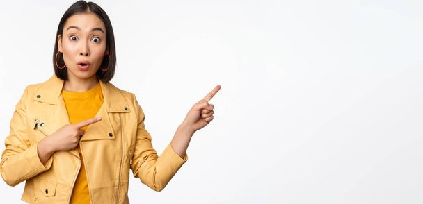 Entusiasta chica asiática señalando con los dedos a la derecha, mostrando publicidad y sonriendo, invitando a la tienda, de pie sobre fondo blanco - Foto, Imagen