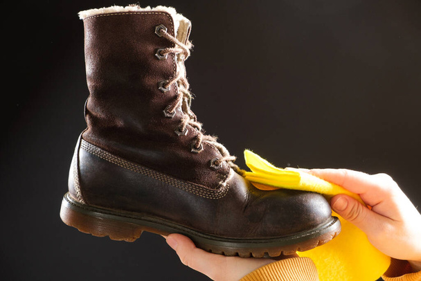Een vrouw reinigt bruine nubuck laarzen op een zwarte achtergrond, veegt het stof met een gele schoen doek. - Foto, afbeelding