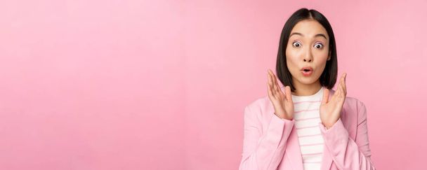 Portrait of asian businesswoman looking surprised at camera, clapping hands and staring excited, smiling, posing against pink background - Fotoğraf, Görsel