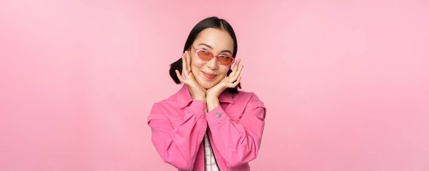 Portrait of stylish cute asian girl, smiling and touching face, looking up dreamy, thoughtful look, standing over pink background - Photo, Image