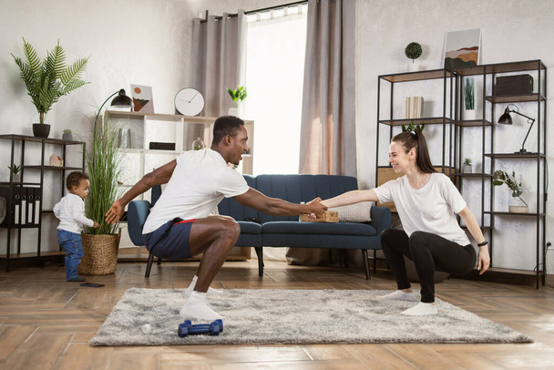 Young multinational multiracial couple doing squats holding hands training together at home - Foto, Bild