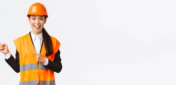 Будівництво, будівництво та промислова концепція. Усміхнена жінка-інженер в шоломі та одязі, що відображає спосіб, вказуючи верхній лівий кут, говорячи про будівництво проекту, білий фон
 - Фото, зображення