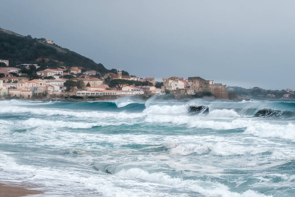 Mares acidentados colidem com a praia em Algajola, na região de Balagne, na Córsega, com aldeia e cidadela à distância - Foto, Imagem