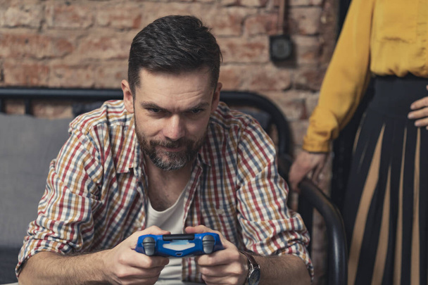 kavkazský muž v rozepnuté kostkované košili hraní her s modrou herní regulátor a ignorování jeho přítelkyně - Fotografie, Obrázek