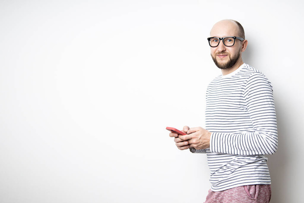Barbudo con gafas sostiene un teléfono en sus manos y se apoya contra una pared blanca. - Foto, imagen