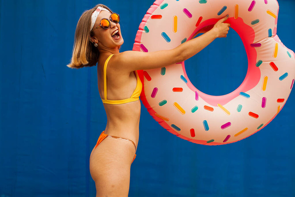 Portret śmiejącej się dziewczyny ubranej w strój kąpielowy posiada nadmuchiwany pierścień odizolowany na niebieskim tle - Zdjęcie, obraz