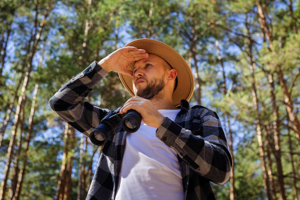 Man tourist in a hat and a plaid shirt looks through binoculars in the forest. - Zdjęcie, obraz