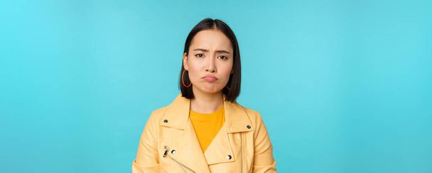 Nahaufnahme eines enttäuschten asiatischen Mädchens, das schmollt, Grimassen schlägt und unzufrieden die Stirn runzelt, launig vor blauem Hintergrund steht - Foto, Bild