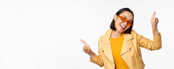 Lächelnde asiatische Brünette mit Sonnenbrille, die Finger seitlich, links und rechts zeigt, Varianten zeigt, lacht und tanzt, Sonnenbrille trägt, weißer Hintergrund - Foto, Bild