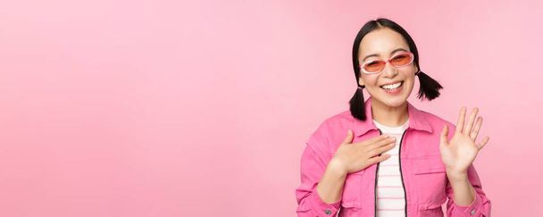 Портрет дружелюбной азиатской девочки-подростка в солнечных очках со словами "привет", машет рукой и улыбается, приветствует вас, привет жест, стоит на розовом фоне - Фото, изображение