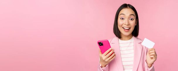 Imagen de la feliz mujer de negocios asiática sonriente mostrando tarjeta de crédito, pagando en línea en la aplicación del teléfono inteligente, orden con teléfono móvil, de pie sobre fondo rosa - Foto, Imagen
